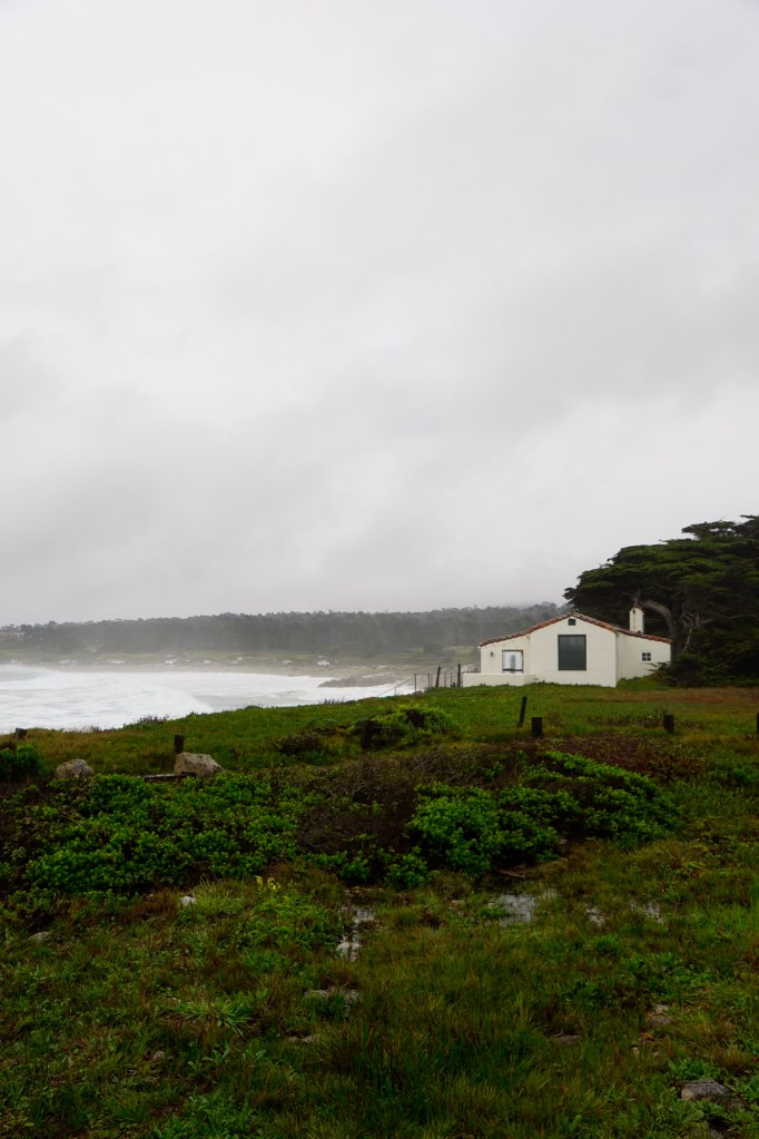 A small white cottage surrounded by green grass and shrubs, set against a grey, overcast sky and a turbulent ocean in the background.